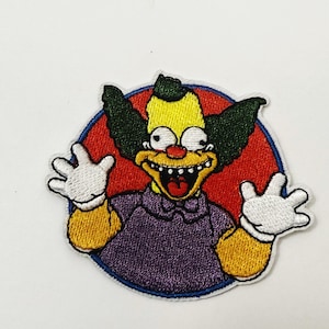 May include: Embroidered patch featuring Krusty the Clown from The Simpsons. The patch shows Krusty with a yellow face, green hair, and a red nose, set against a red circle. He wears a purple shirt and white gloves.