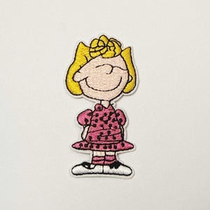 May include: Embroidered patch featuring Sally Brown from the Peanuts comic strip. The patch shows Sally in a pink dress with black polka dots, yellow hair, and a smiling expression. The patch has a white border.
