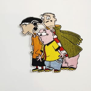 Peut inclure: Écusson brodé représentant trois personnages de la série animée "Ed, Edd n Eddy". L'écusson présente les personnages dans des couleurs vives, avec des coutures détaillées. Les personnages sont debout, rapprochés, l'un d'eux portant une cape verte.