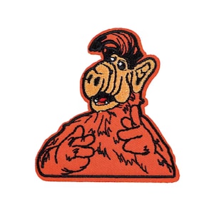 May include: Embroidered patch featuring the character ALF from the 1980s sitcom. The patch has a red-orange background with a black border. ALF is depicted with orange fur, a tan face, and black hair. His mouth is open, and he is giving a thumbs-up.