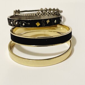 Op de afbeelding: Een stapel van vier armbanden in verschillende ontwerpen. De bovenste armband is zilverkleurig met een herhalend ruitpatroon. Daaronder bevindt zich een zwarte armband met gouden en zilveren accenten. De volgende armband is goudkleurig met een zwarte band.