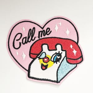 Könnte beinhalten: Gestickter Aufnäher mit einem Cartoon-Telefon-Design, das einen roten Hörer und ein zwinkerndes Auge zeigt. Der Aufnäher befindet sich auf einem rosa herzförmigen Hintergrund mit den Worten "Call me" in schwarzer Schrift. Weiße Sternausbrüche sorgen für einen Hauch von Glanz.