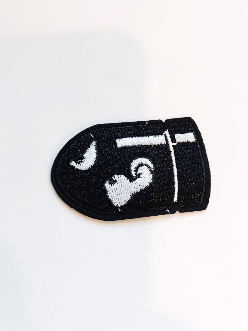 Puede incluir: Parche bordado negro con forma de bala con un contorno blanco y un coraz&oacute;n blanco en el interior.
