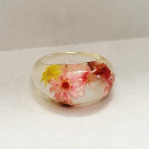Puede incluir: Un anillo de resina transparente con flores secas incrustadas en tonos rosa, amarillo y rojo. El anillo tiene una banda ancha y un acabado liso y pulido. Las flores son visibles a través de la resina transparente.