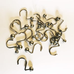May include: A collection of shiny, silver-colored shower curtain hooks. Each hook has a curved design with a small, round ball at the end of the hook. The hooks are scattered on a white surface.