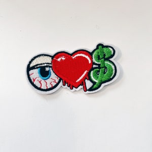 May include: An embroidered patch with a red heart, a green dollar sign, and a blue eye with red blood dripping from the heart.