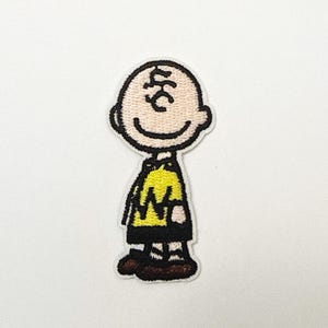 May include: Embroidered patch of Charlie Brown from Peanuts. The patch depicts Charlie Brown in his yellow shirt with a black zig-zag, black shorts, and brown shoes. It has a white border.