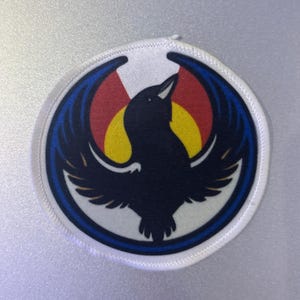 May include: Round patch with a black bird design, wings spread, set against a yellow sun, red, and blue elements. The patch has a white border and is likely an emblem or logo.