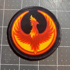 May include: Round black patch featuring a stylized phoenix design. The phoenix is depicted in shades of red, orange, and yellow, with a black background. The patch has a stitched border.