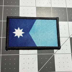 Puede incluir: Parche rectangular con un diseño azul y celeste. Una estrella blanca de ocho puntas está en el lado izquierdo del parche. El parche está bordeado con costuras negras.