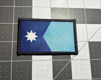May include: Rectangular patch with a blue and light blue design. A white eight-pointed star is on the left side of the patch. The patch is bordered with black stitching.