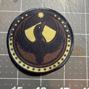 May include: Round patch with a bird emblem in black, brown, and gold. The design includes a star and a ring of stars around the bird. Suitable for attaching to clothing or bags.