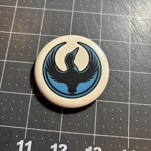 May include: A round button with a cream-colored background. The button features a black bird design with blue accents. The bird is stylized with outstretched wings. The button is on a black cutting mat with white grid lines.