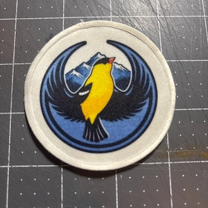 May include: Round patch with a bird design. The bird is yellow with black wings and a red beak, set against a blue background with mountain imagery. The patch has a white border.