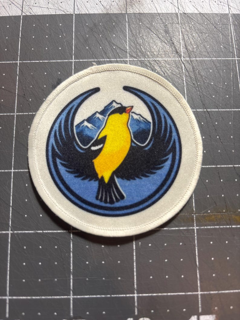 Op de afbeelding: Ronde patch met een vogelontwerp. De vogel is geel met zwarte vleugels en een rode snavel, tegen een blauwe achtergrond met berglandschap. De patch heeft een witte rand.