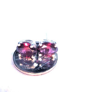 May include: Two oval-cut, deep red garnet stud earrings set in silver-toned metal prongs. The earrings are positioned on top of a United States dime coin, which serves as a scale reference for the size of the faceted gemstones.