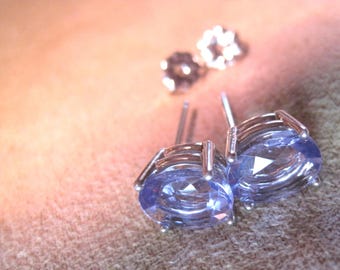 May include: A pair of stud earrings with oval, light blue gemstones set in silver metal. Two additional silver earrings are visible in the background. The earrings are displayed on a neutral surface.