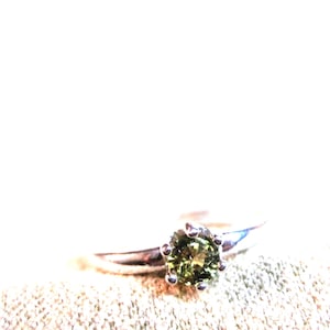 May include: A silver-toned ring featuring a round, faceted olive-green gemstone held in a six-prong setting. The band is smooth and metallic, tapering slightly as it extends from the central stone, which displays a vibrant, earthy green hue.