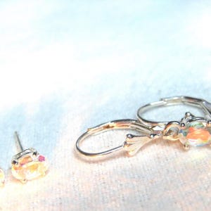 May include: A set of jewelry consisting of two stud earrings and two leverback earrings. Each earring features a small, round, iridescent stone with a metallic setting that reflects light with rainbow hues, creating a shimmering effect.