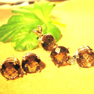 May include: A jewelry set featuring a pendant and two pairs of earrings with large, faceted, brown gemstones. The jewelry is set in a gold-colored metal. A green leaf is in the background, set against a yellow surface.