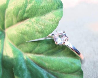 May include: A silver ring with a clear, round gemstone set in a prong setting. The ring is resting on a green leaf.