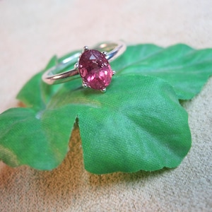 May include: A silver ring with a pink oval gemstone. The ring is set on a green leaf, with a small pink gemstone visible to the right. The ring's band is thin and polished, and the gemstone is held in place by four prongs.