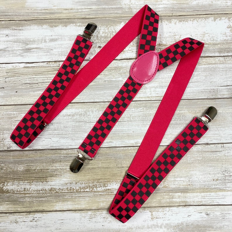 May include: Red suspenders with black and white checkered pattern. The suspenders have silver clips and a pink leather button.