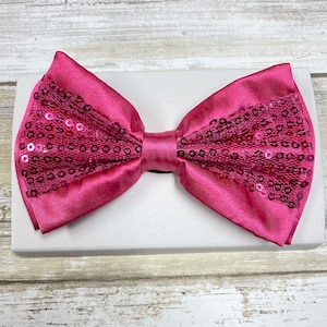 May include: A pink bow tie with sequins. The bow tie is made of satin and has a layer of sequins on top. The sequins are arranged in a pattern that resembles a net.