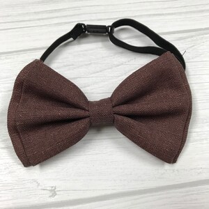 May include: A brown bow tie with an adjustable elastic strap. The bow tie is made of a textured fabric.