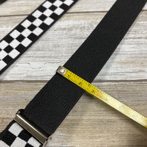 May include: Black elastic suspenders with a checkered pattern. The suspenders are measured with a tape measure, showing they are 1 3/4 inches wide.