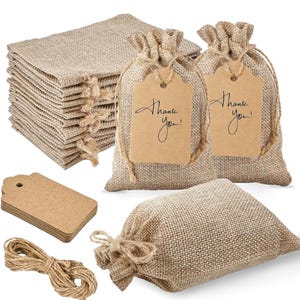 Peut inclure: Ensemble de sacs cadeaux en toile de jute beige, étiquettes et ficelle. Les sacs ont une fermeture à cordon. Deux sacs portent des étiquettes avec l'inscription "Thank you!". Une pile d'étiquettes et une bobine de ficelle sont également incluses.