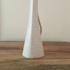 May include: A white ceramic vase with a textured surface and a unique, asymmetrical shape. The vase has a tall, triangular body that tapers upwards to a slanted opening. A small handle curves from the side. The vase is set on a wooden surface.