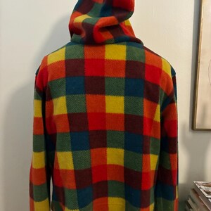 May include: A cropped fleece jacket with a hood, featuring a vibrant plaid pattern in red, orange, yellow, green, and blue. The jacket has long sleeves and a rounded hemline. The hood is also plaid.