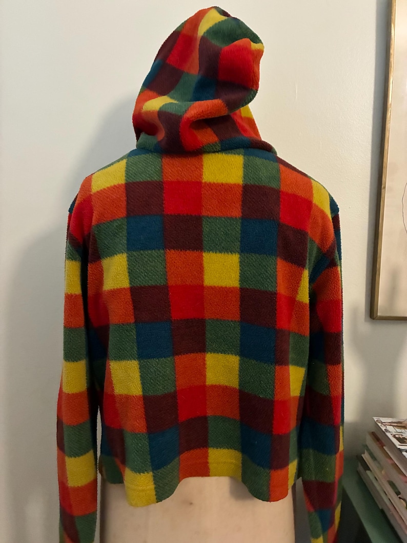 May include: A cropped fleece jacket with a hood, featuring a vibrant plaid pattern in red, orange, yellow, green, and blue. The jacket has long sleeves and a rounded hemline. The hood is also plaid.