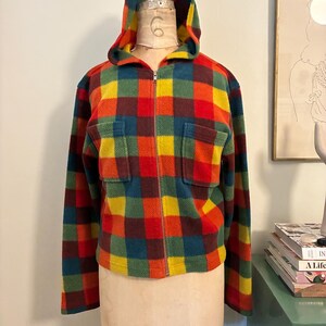 May include: A colorful, hooded fleece jacket with a plaid pattern in red, orange, yellow, green, and blue. The jacket has a full zipper, two front pockets, and a hood. The jacket is displayed on a mannequin.