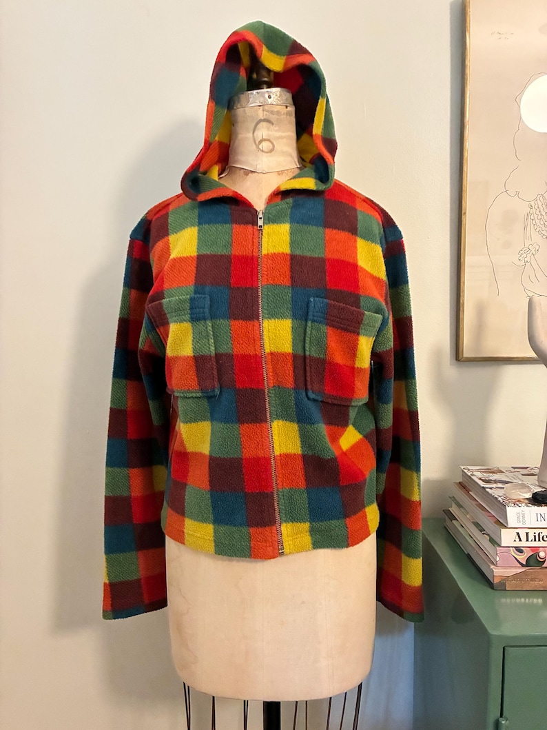May include: A colorful, hooded fleece jacket with a plaid pattern in red, orange, yellow, green, and blue. The jacket has a full zipper, two front pockets, and a hood. The jacket is displayed on a mannequin.