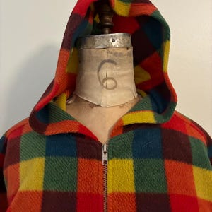May include: A hooded fleece jacket in a vibrant plaid pattern of red, orange, yellow, green, and blue squares. The jacket features a full zipper and a cozy, warm design. The hood is lined with a contrasting yellow fabric.
