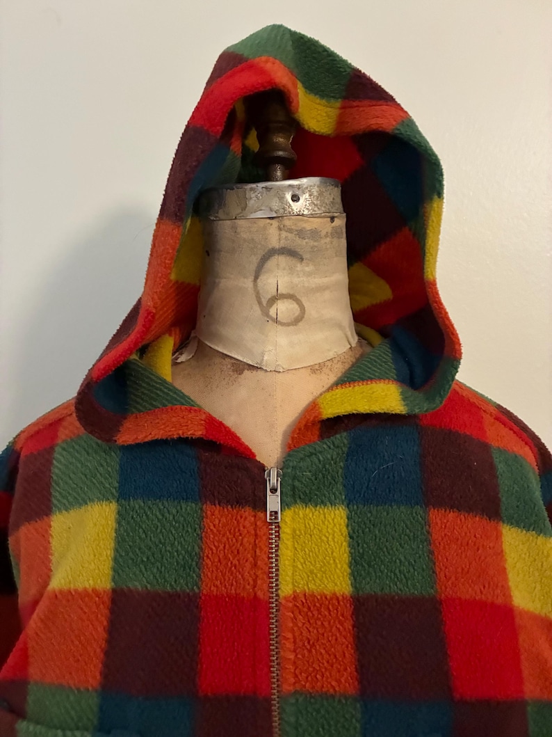 May include: A hooded fleece jacket in a vibrant plaid pattern of red, orange, yellow, green, and blue squares. The jacket features a full zipper and a cozy, warm design. The hood is lined with a contrasting yellow fabric.