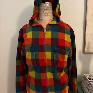 May include: A colorful, plaid fleece zip-up hooded jacket. The jacket features a vibrant pattern of red, orange, yellow, green, and blue squares. It has a full zipper, two front pockets, and a hood. The jacket is displayed on a mannequin.