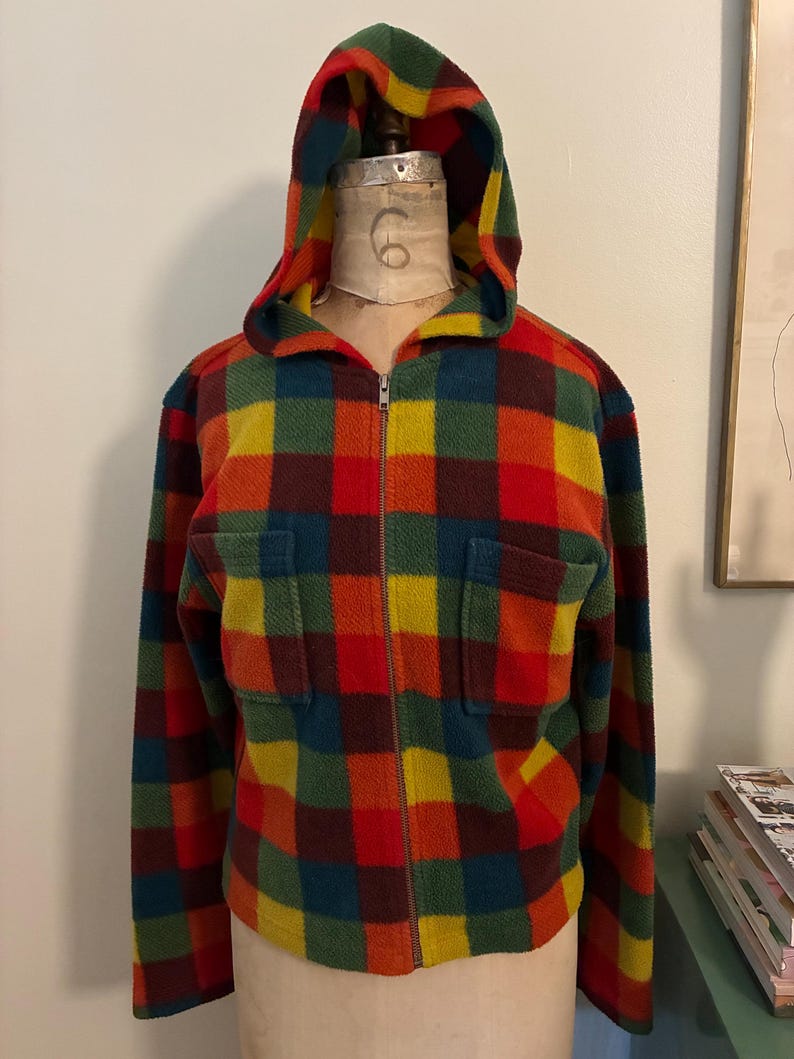 May include: A colorful, plaid fleece zip-up hooded jacket. The jacket features a vibrant pattern of red, orange, yellow, green, and blue squares. It has a full zipper, two front pockets, and a hood. The jacket is displayed on a mannequin.