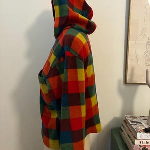 May include: A hooded jacket with a vibrant plaid pattern in red, orange, yellow, green, and blue. The jacket features a large hood and a front pocket. The fabric appears soft and warm, suitable for cooler weather.