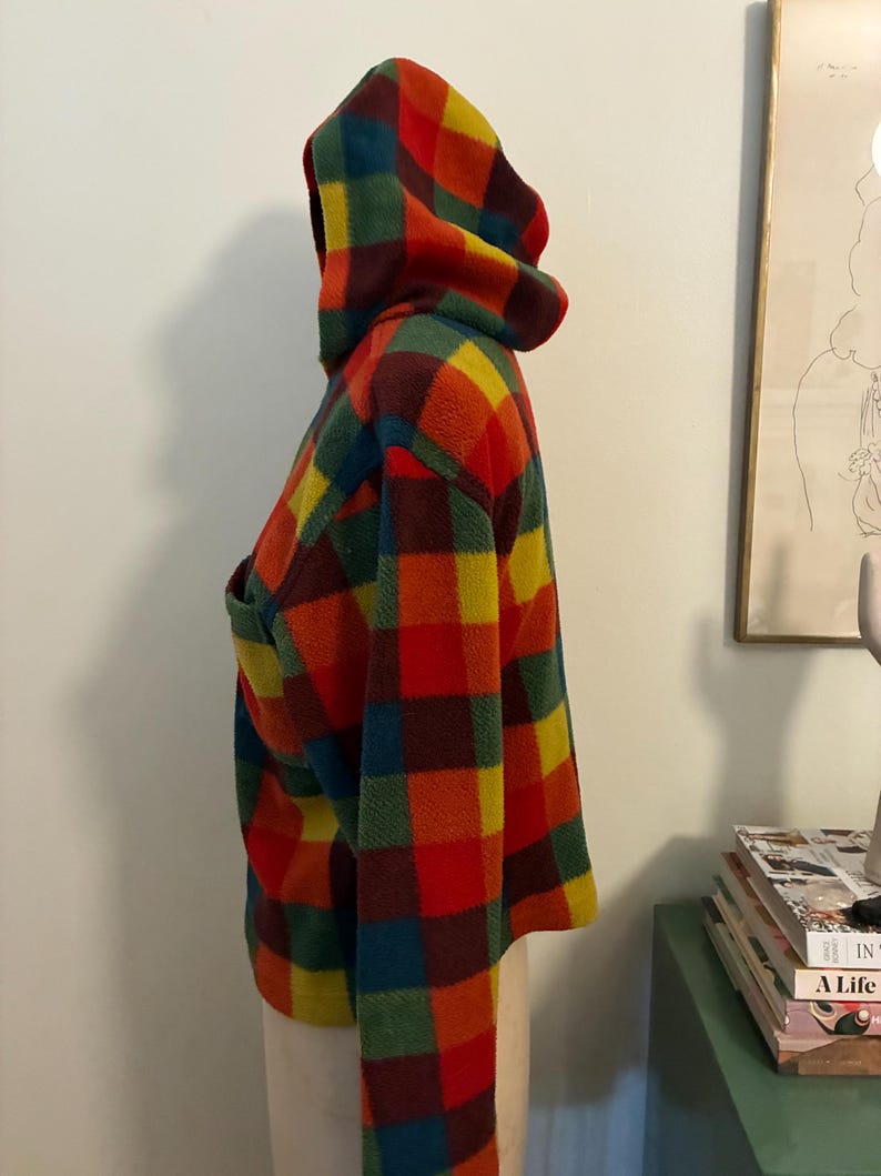 May include: A hooded jacket with a vibrant plaid pattern in red, orange, yellow, green, and blue. The jacket features a large hood and a front pocket. The fabric appears soft and warm, suitable for cooler weather.