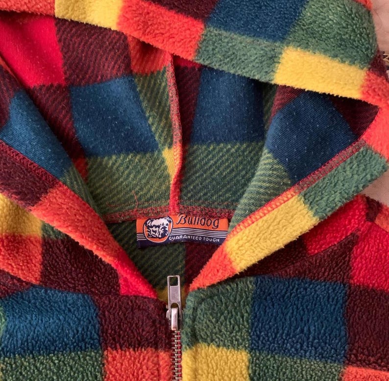 May include: A colorful, hooded fleece jacket with a plaid pattern in shades of red, orange, yellow, green, and blue. The jacket has a full zipper and a Bulldog brand label. The fleece material appears soft and warm.