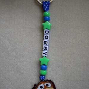 May include: A keyring featuring a blue ribbon with white polka dots, green and blue beads, and two green star-shaped beads. White square beads spell out the name BOBBY. A metal charm of a brown and tan dog face hangs at the bottom of the ribbon.