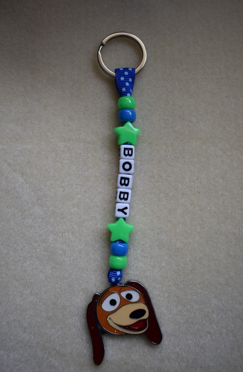 May include: A keyring featuring a blue ribbon with white polka dots, green and blue beads, and two green star-shaped beads. White square beads spell out the name BOBBY. A metal charm of a brown and tan dog face hangs at the bottom of the ribbon.