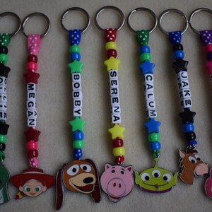 May include: A collection of twelve personalised keychains featuring colourful plastic beads, star-shaped spacers, and white letter cubes spelling names like Jacob, Lukas, Ollie, Megan, Bobby, Serena, Calum, Jake, Leanna, George, and Aimee, with character charms.