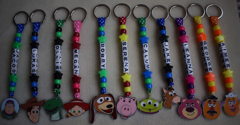 May include: A collection of twelve personalised keychains featuring colourful plastic beads, star-shaped spacers, and white letter cubes spelling names like Jacob, Lukas, Ollie, Megan, Bobby, Serena, Calum, Jake, Leanna, George, and Aimee, with character charms.