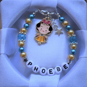 May include: A blue and gold beaded bracelet with a charm of Snow White and a star charm. The bracelet has the name "PHOEBE" spelled out in white letters.