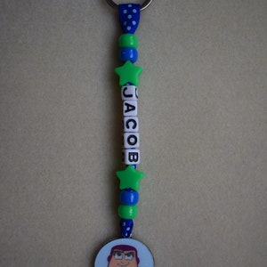 May include: A keyring featuring a blue ribbon with white polka dots, green and blue spherical beads, two green star-shaped beads, and white cube beads spelling the name JACOB. A circular charm depicting a cartoon space ranger character is attached.