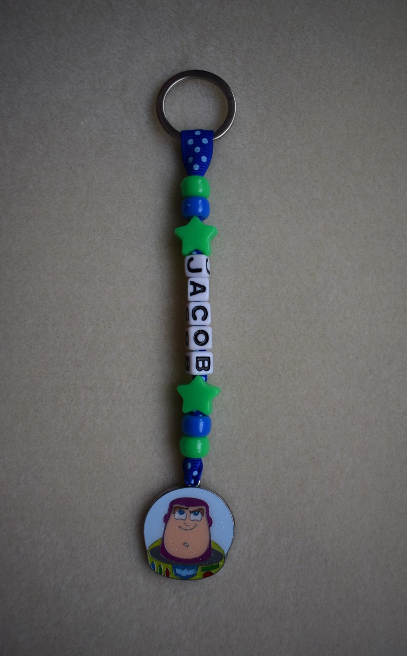 May include: A keyring featuring a blue ribbon with white polka dots, green and blue spherical beads, two green star-shaped beads, and white cube beads spelling the name JACOB. A circular charm depicting a cartoon space ranger character is attached.